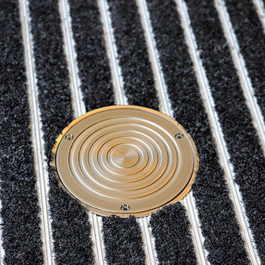 A bronze-colored drain inspection point with anti-slip grooves on the lid, set against a striped background.